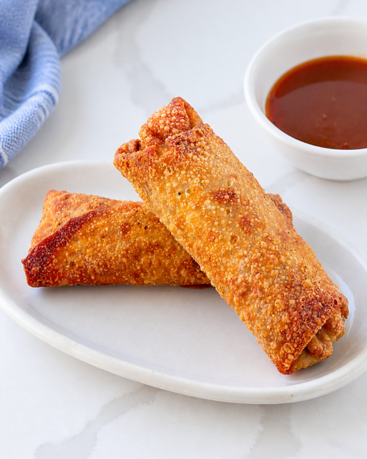 Two crisp homemade pork egg rolls on an ivory platter with sweet and sour sauce and a softly bunched blue striped linen.