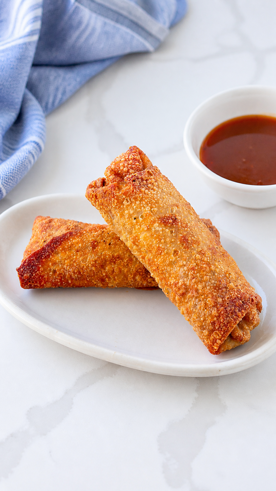 Two crisp homemade pork egg rolls on an ivory platter with sweet and sour sauce and a softly bunched blue striped linen.