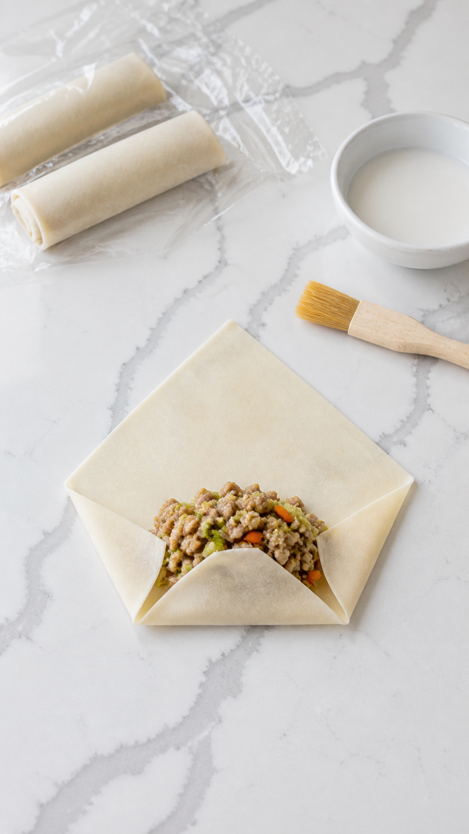 An egg roll wrapper folded around pork and vegetable filling with slurry ready for sealing.