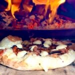 Wood-fired pizza baking beside bright flames inside an Ooni Karu 2 Pro Multi-Fuel Pizza Oven.