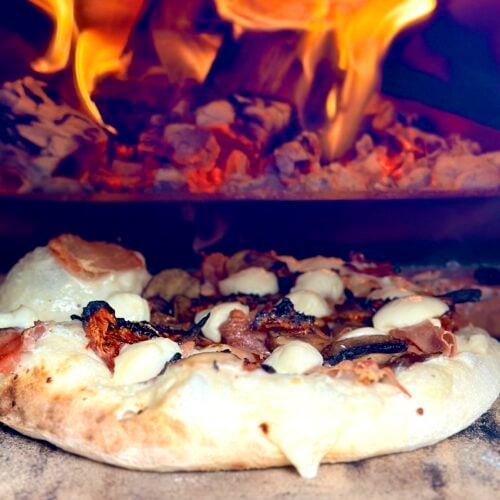 Wood-fired pizza baking beside bright flames inside an Ooni Karu 2 Pro Multi-Fuel Pizza Oven.