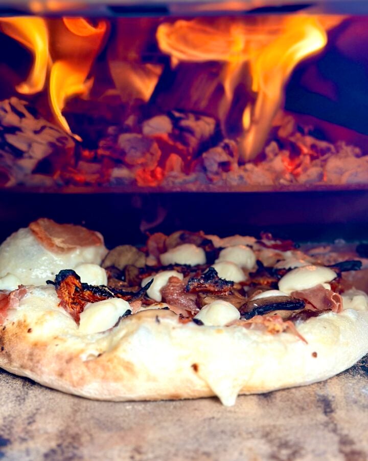 Wood-fired pizza baking beside bright flames inside an Ooni Karu 2 Pro Multi-Fuel Pizza Oven.