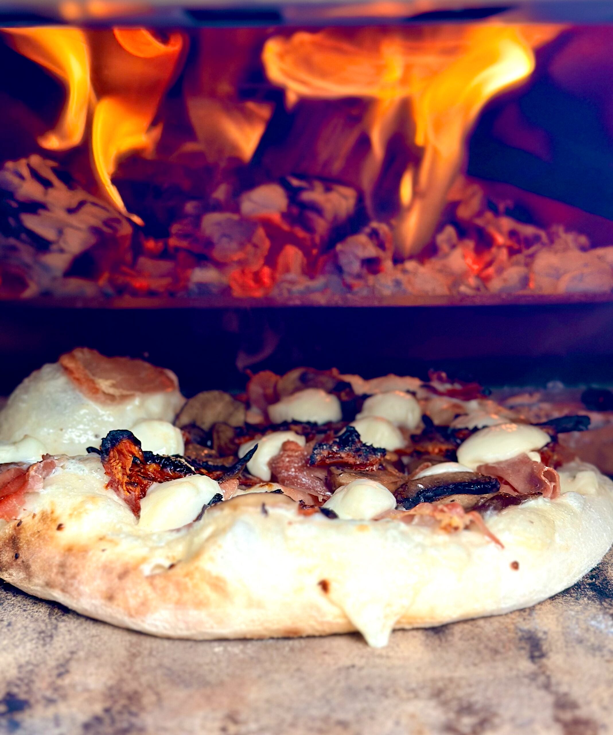Wood-fired pizza baking beside bright flames inside an Ooni Karu 2 Pro Multi-Fuel Pizza Oven.