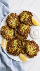 Crispy zucchini fritters arranged on a white plate with lemon dill sauce and lemon wedges.