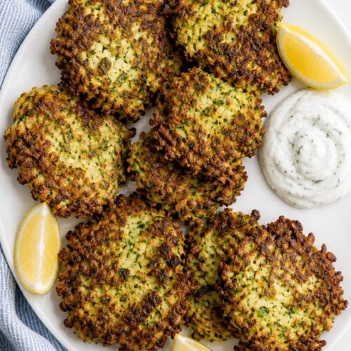 Crispy zucchini fritters arranged on a white plate with lemon dill sauce and lemon wedges.