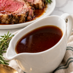 Dark red wine au jus in a white gravy boat beside sliced prime rib.