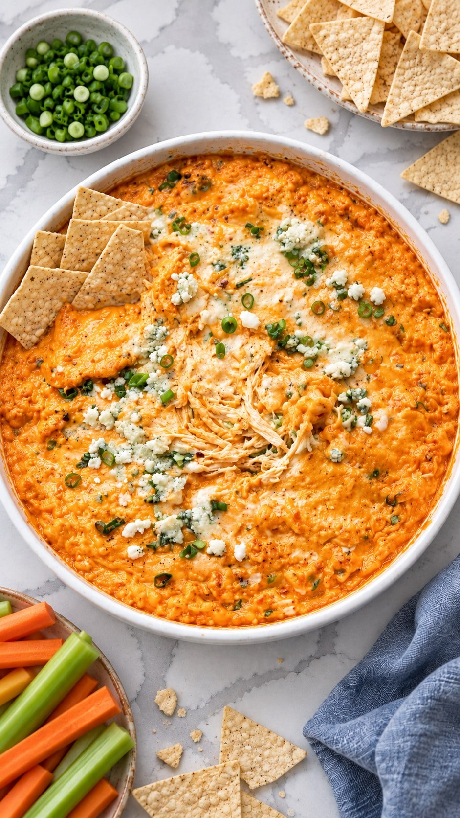 Buffalo chicken dip in a round white serving dish with tortilla chips, blue cheese crumbles, and green onions.