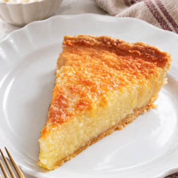 Buttermilk pie slice with a golden top and creamy filling, on a scalloped white plate beside coffee and whipped cream