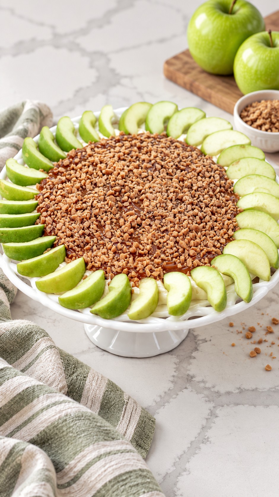Caramel apple dip topped with toffee and surrounded by Granny Smith wedges on a white pedestal platter