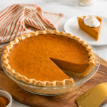 Classic pumpkin pie with one wedge removed and a plated slice with whipped cream nearby.