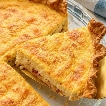 A creamy bacon and Gruyère Quiche Lorraine wedge with a naturally uneven cut and flaky crumbs on a gold pie server.