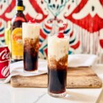 Colorado Bulldog with a dark coffee-and-cola base, ivory cream swirls, ice and a red cherry against a vibrant red and teal patterned background.