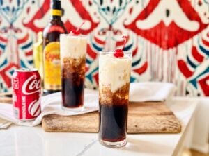 Colorado Bulldog with a dark coffee-and-cola base, ivory cream swirls, ice and a red cherry against a vibrant red and teal patterned background.