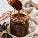 A gold spoon lifts speckled homemade vanilla bean paste above a glass jar styled with linen and vanilla pods.