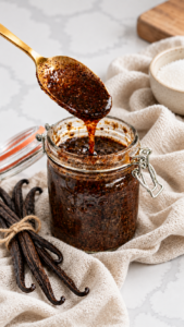 A gold spoon lifts speckled homemade vanilla bean paste above a glass jar styled with linen and vanilla pods.