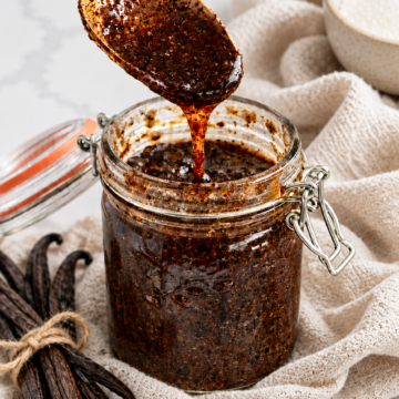 A gold spoon lifts speckled homemade vanilla bean paste above a glass jar styled with linen and vanilla pods.