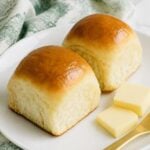 Two soft golden homemade yeast rolls served on a white plate with butter.