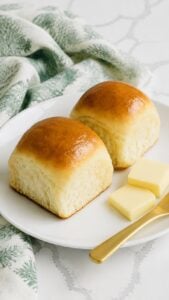 Two glossy golden homemade yeast rolls on a white plate with butter.