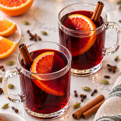 Mulled wine with orange slices and cinnamon in clear handled mugs, surrounded by whole spices.