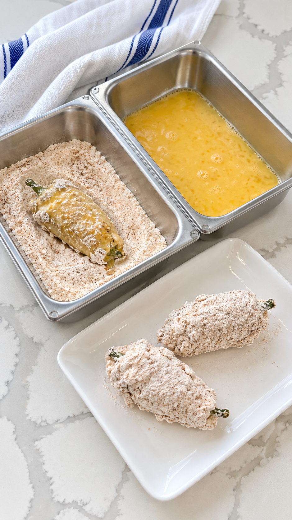 Stuffed jalapeños coated in seasoned flour beside egg wash and flour trays.