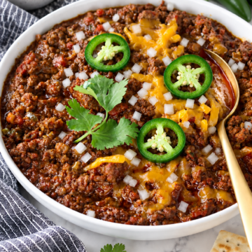 Texas chili con carne topped with cheddar, jalapeño slices, finely diced white onion and cilantro in a white bowl.