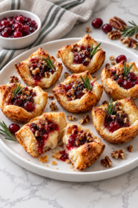Cranberry Brie Bites with golden puff pastry, cranberry sauce, pecans, and rosemary on a white platter.