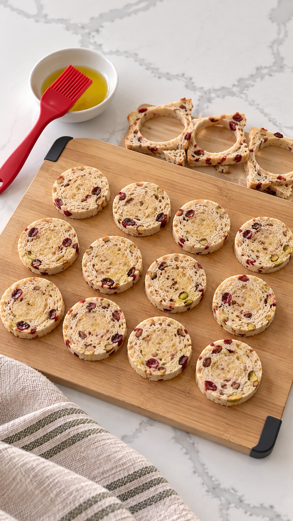 Twelve untoasted cranberry-pistachio bread rounds with reserved bread scraps and olive oil.