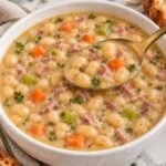 Ham and bean soup with navy beans, diced ham, carrots and celery in a white bowl, with a spoonful lifted.