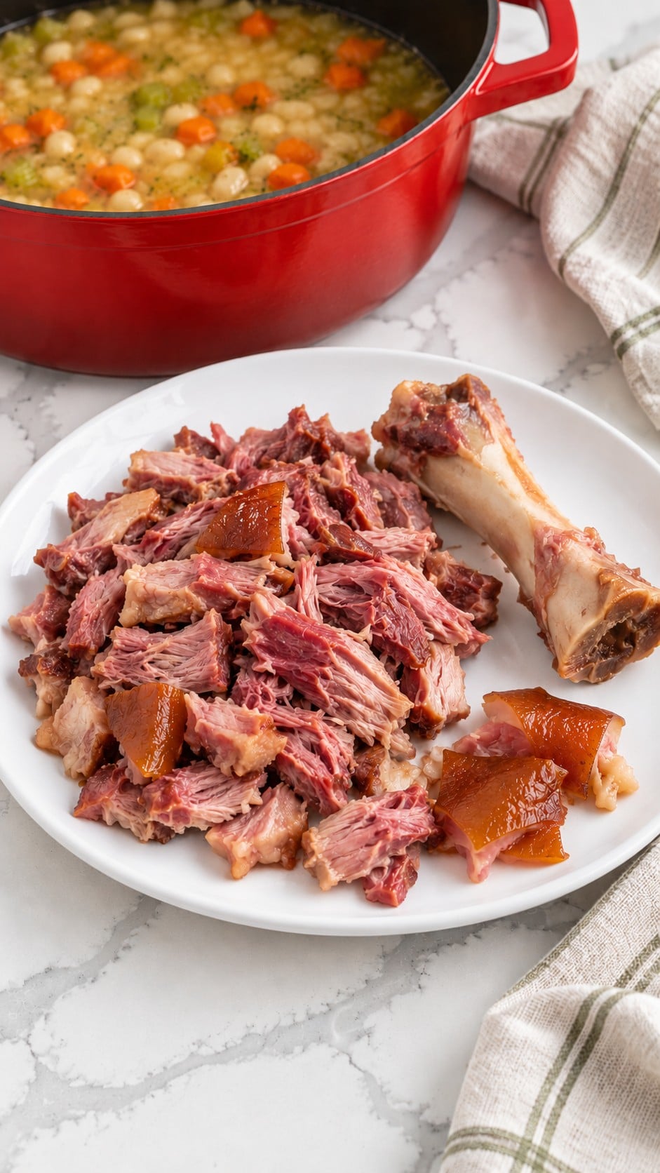 Cooked meat pulled from the ham hock, with bone and skin separated on a white plate.